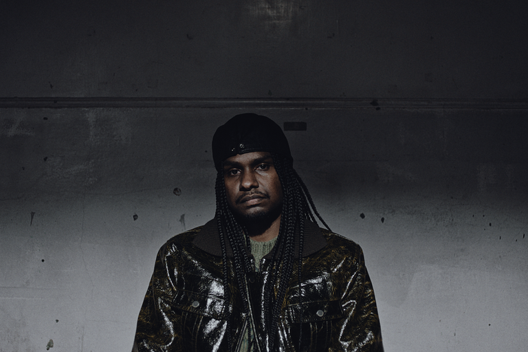 Indigenous Australian rapper Baker Boy posing in a textured jacket and cap against a dark wall in a moody promo portrait.